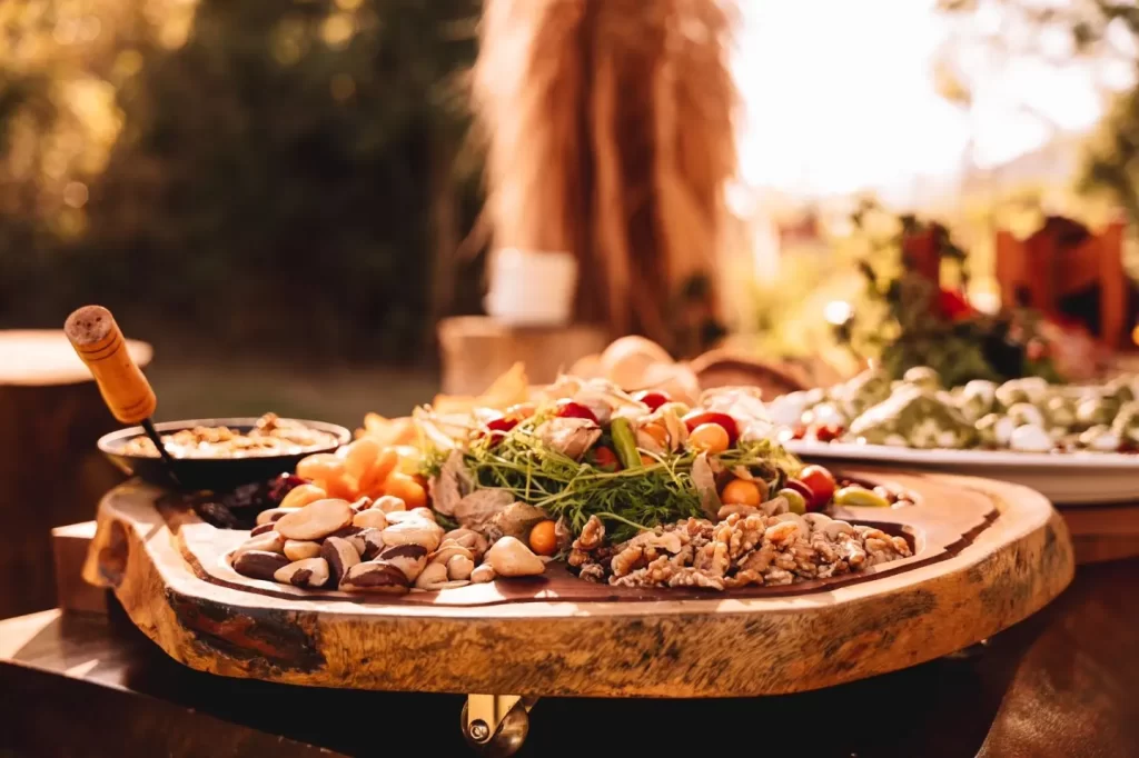 Fotografia gastronômica horizontal de tábua rústica circular de madeira repleta de comidas típicas da mata atlântica como castanhas, ramos de ervas verdes aromáticas. Segundo plano desfocado mostra travessas adicionais com outros tipos de comida. Cenário ao ar livre banhado por luz dourada quente, com fundo desfocado criando bokeh natural.
